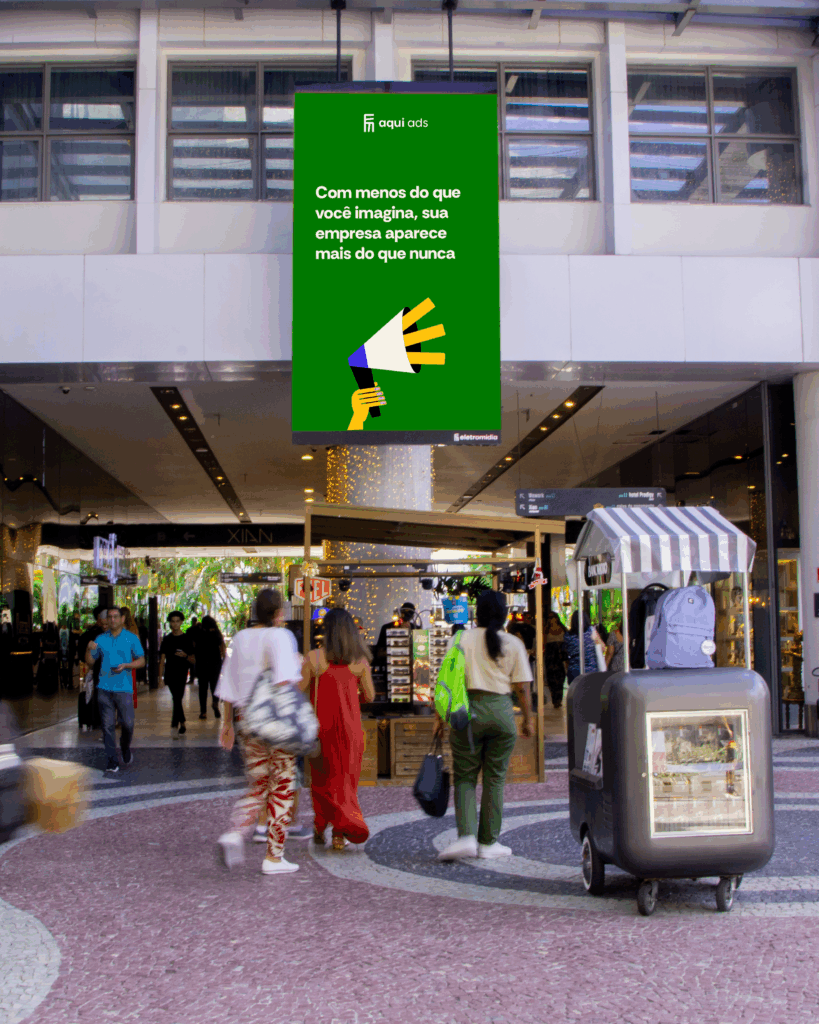 Pessoas caminhando em um shopping center moderno com um anúncio digital suspenso do Aqui Ads. O anúncio verde exibe a frase 'Com menos do que você imagina, sua empresa aparece mais do que nunca' e uma ilustração de um megafone.