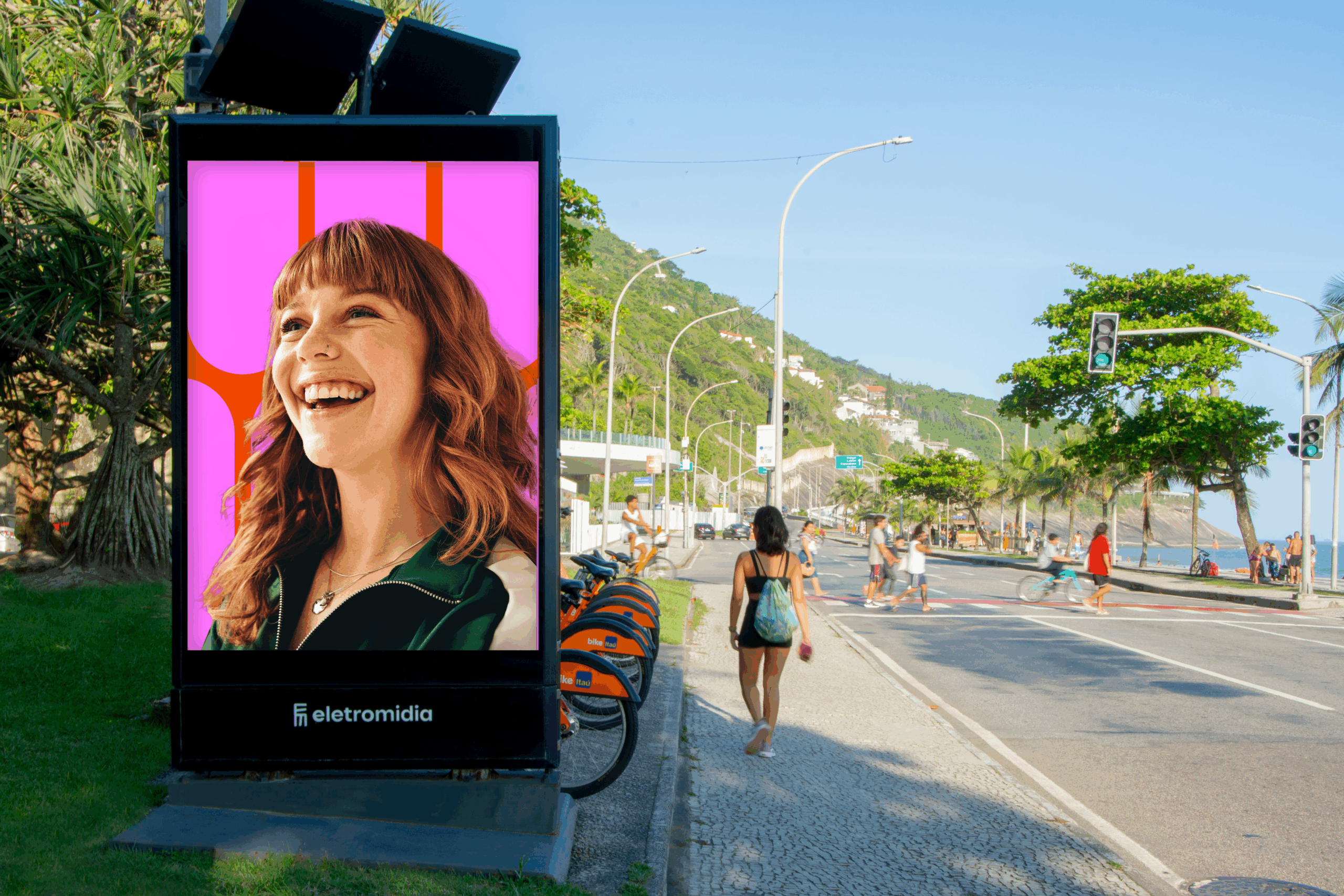 Painel de publicidade digital da Eletromidia exibindo um anúncio vibrante com uma mulher sorrindo em um fundo rosa, localizado ao lado de uma estação de bicicletas Tembici em uma rua movimentada do Rio de Janeiro, com praia e montanhas ao fundo.